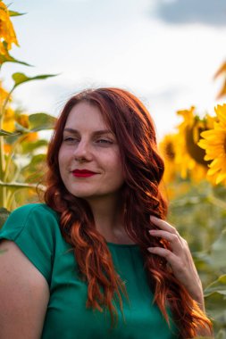 Dudaklarında kırmızı ruj ve yaz ayçiçeği tarlasında poz veren yeşil elbiseli kızıl saçlı kıvırcık kız.