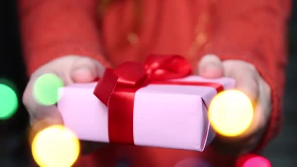 Female hands hold surprise gift box with red ribbon. Blurred garland in the foreground. Happy New Year, Birthday, Christmas, Womens Day or ValentineDay. — Stock Video