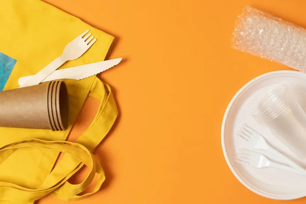 Plastic cup, plate, forks, paper cups, natural cotton yellow bag, forks and knives on an orange background. Confrontation. Ecological catastrophy