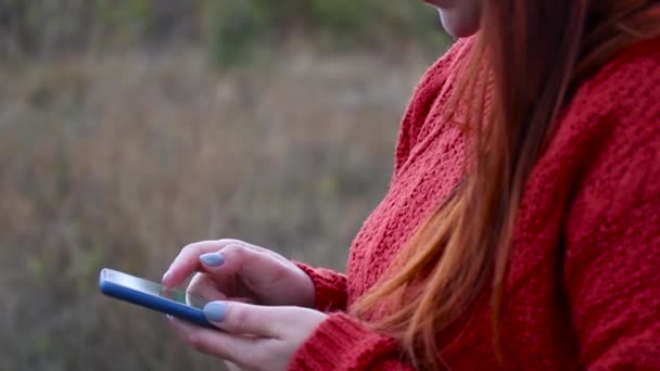 Τα κοριτσίστικα χέρια κρατάνε ένα smartphone. Ένα νεαρό κορίτσι με ένα κόκκινο πουλόβερ χρησιμοποιεί ένα τηλέφωνο ενώ στέκεται στο δρόμο. — Αρχείο Βίντεο