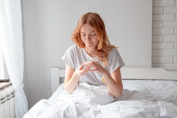 年轻的金发姑娘抱着面霜坐在床上的毛毯下. — 图库照片