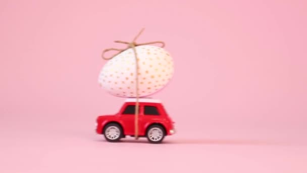 Маленькая красная игрушечная машинка с золотым яйцом на капоте на розовом фоне. Доставка подарков. Концепция счастливой еды — стоковое видео
