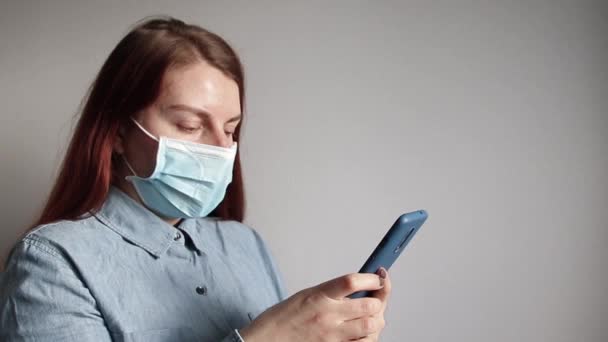 En ung flicka i en medicinsk mask talar på en smartphone mot en grå vägg. — Stockvideo