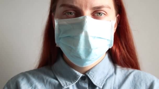 Молодая девушка с рыжими волосами и зелеными глазами в медицинской маске смотрит в камеру. Защита от вирусов, экологическая катастрофа и загрязнение воздуха — стоковое видео