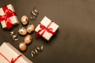 Koyu bir arka planda sürpriz hediye kutularıyla altın yumurtalar ve mesaj yeri. Kırmızı kurdele süsleme. Bayramlık paskalya kompozisyonu