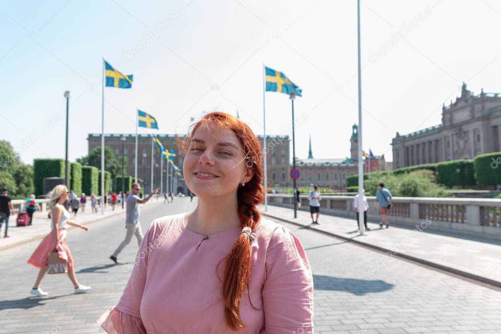 Cute red haired girl with a braid hairstyle in powdery color dress looks at the camera on sunny day outdoors. Stogkolm, Sweden