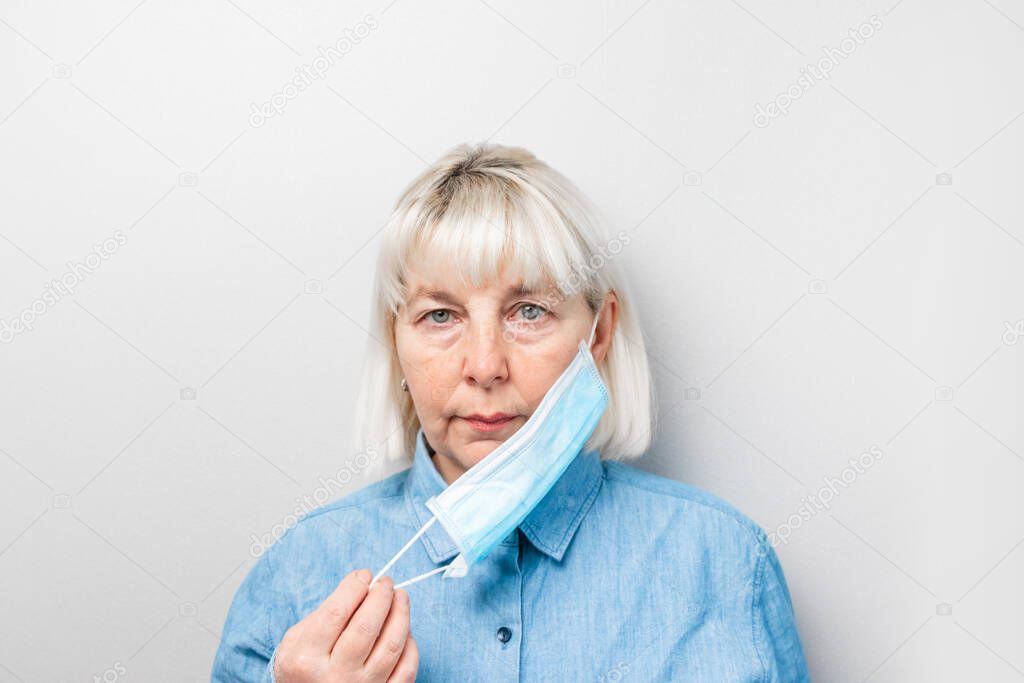 Adult blond woman in a blue shirt puts on a medical mask against a gray wall. Protection against viruses, germs, pneumonia epidemic