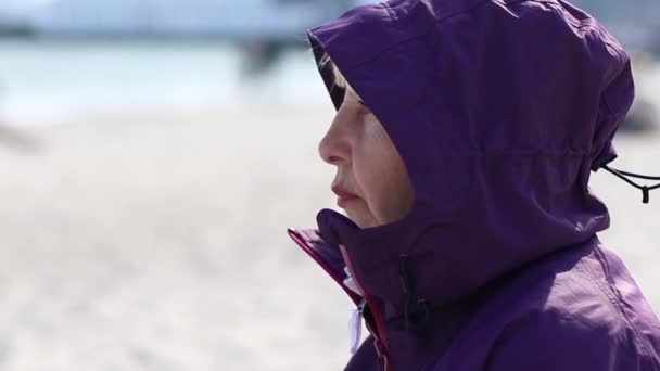 Vuxen kvinna i lila jacka och huva äter en smörgås sittande på stranden nära havet en solig dag, på våren — Stockvideo