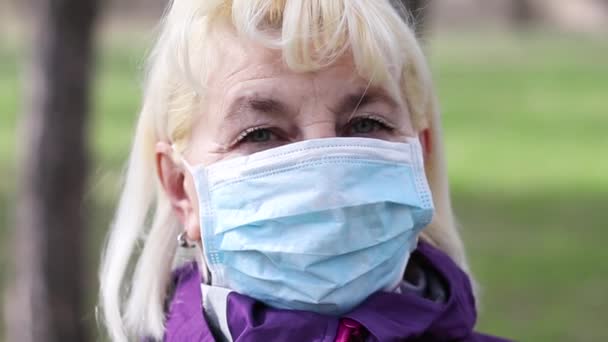 Dospělá blondýna v lékařské masce se dívá na fotoaparát na rozmazaném pozadí mimo dům — Stock video