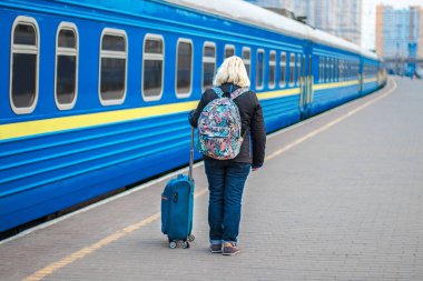 Siyah ceketli yetişkin sarışın kadın tren istasyonunun peronuna döner, güneşli bir günde trenin gelişini bekler.