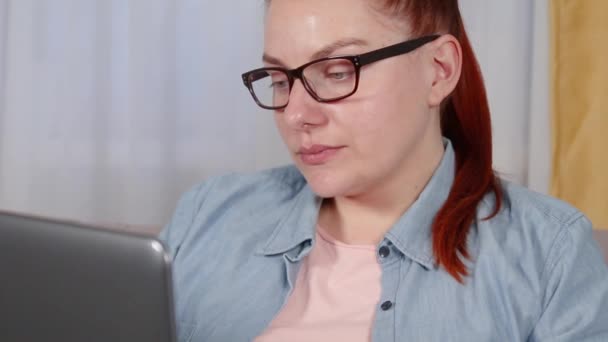 Mulher bonita em óculos e uma camisa trabalhando em um laptop, sentado na sala de estar. bocejo e cansado — Vídeo de Stock