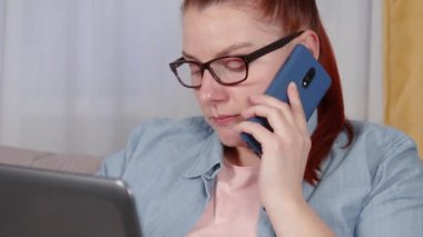 Oturma odasında dizüstü bilgisayarla çalışan yorgun genç bir kadının portresi. Cep telefonuyla konuşan bir kadın..