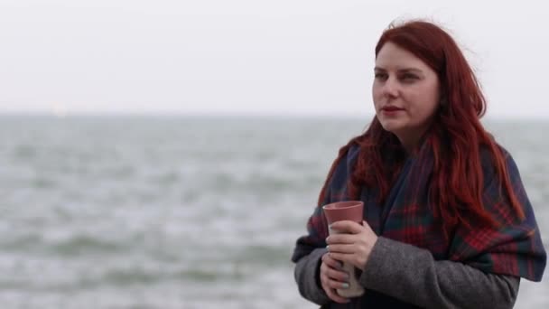 Chica joven con el pelo largo y rojo, con un abrigo cálido y bufanda de lana, sostiene un termo de metal con una bebida caliente en el mar. El cabello se desarrolla en el aire — Vídeo de stock