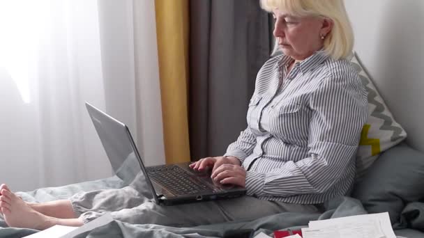 Volwassen vrouw in een gestreept hemd en huisbroek werkend op een laptop zittend in de slaapkamer thuis. Werk thuis concept. Werk vanuit huis concept — Stockvideo