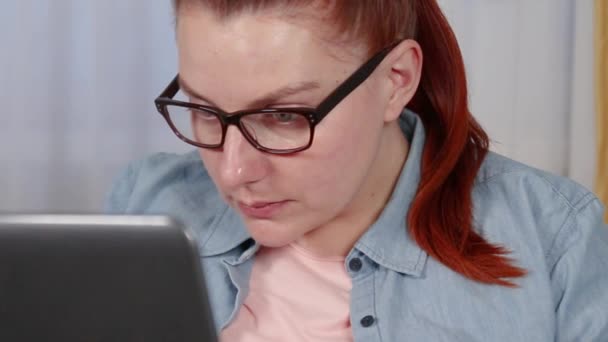 Όμορφη νεαρή γυναίκα με γυαλιά και ένα πουκάμισο που δουλεύει σε ένα φορητό υπολογιστή που κάθεται σε ένα σαλόνι — Αρχείο Βίντεο
