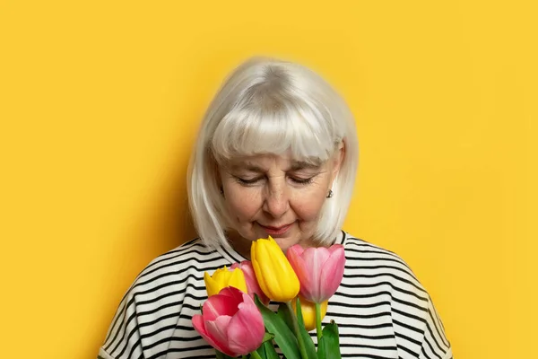 Carino sorridente donna bionda adulta in un maglione a righe tiene un mazzo di fiori di tulipani e li annusa. Giornata internazionale della donna. Buona festa della mamma . — Foto Stock