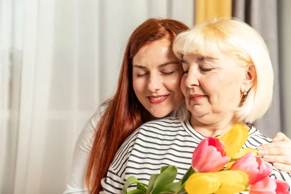 Día Internacional de la Mujer. Feliz Día de las Madres. Mujer rubia adulta sostiene un ramo de tulipanes y su hija abraza la casa. Generación de mujeres —  Fotos de Stock