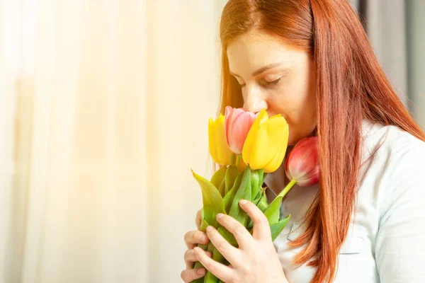 Dia das Mulheres 8 de março. Jovem alegre alegre com cabelo longo vermelho segura um lindo buquê de tulipas de flores e cheira no quarto — Fotografia de Stock