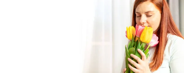 Krásná mladá dívka s červenými dlouhými vlasy drží krásnou kytici květin tulipánů a čichá k nim v místnosti — Stock fotografie