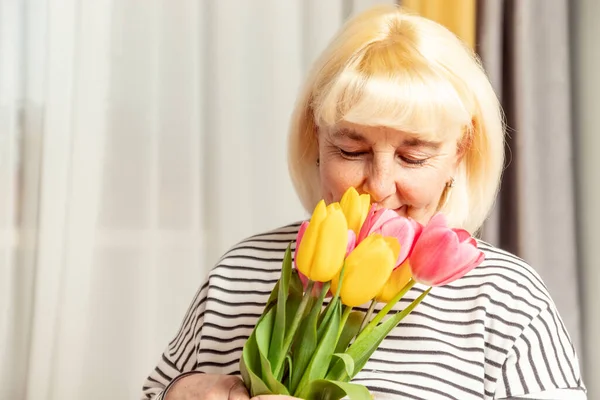 Feliz día de las madres. Mujer rubia adulta oliendo un ramo de tulipanes flores sobre un fondo gris —  Fotos de Stock