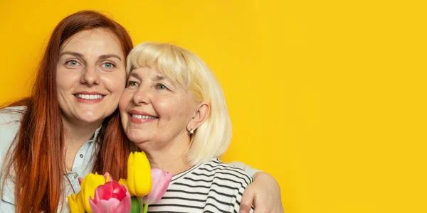 Mulher adulta e sua filha menina abraçou e segura em suas mãos um buquê de tulipas em um fundo de parede amarelo. Dia das Mães — Fotografia de Stock