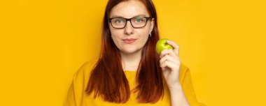 Yeşil elma yiyen güzel bir kız, sarı bir duvarın arkasında duruyor. Diyet ve uygun beslenme konsepti.