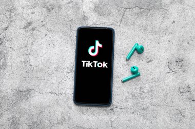 Akıllı telefon ekranında videolu Tiktok uygulaması ve gri arka planda kablosuz kulaklık