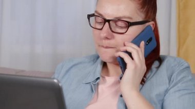 Oturma odasında dizüstü bilgisayarla çalışan yorgun genç bir kadının portresi. Cep telefonuyla konuşan bir kadın..