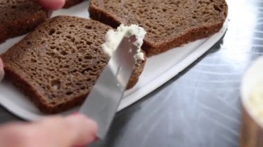 Çok tahıllı ekmeğin üzerinde krem peynirli bıçak lekesi olan bir kadın eli. Kapat.