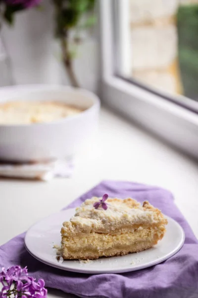 Кусок вкусного чизкейка на белой тарелке на подоконнике, на заднем плане букет сирени, вертикальное фото — стоковое фото