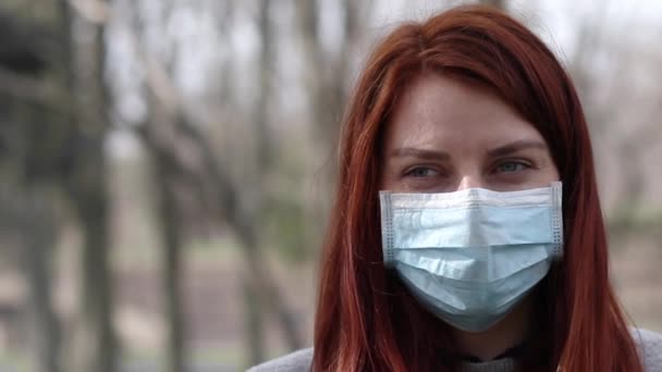 Covid-19 epidemie. Een jong rood haarmeisje met een medisch masker kijkt naar de camera in het park. Wazige achtergrond. — Stockvideo