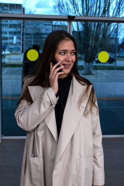 Genç, kahverengi saçlı, yeşil gözlü ve açık renkli siperli güzel bir kız dükkanın önünde konuşuyor.