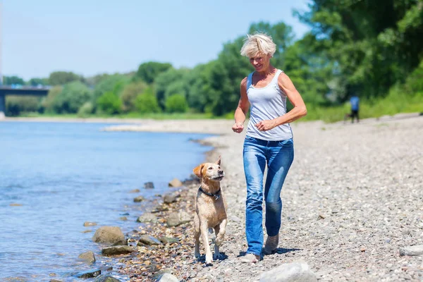 Rijpe vrouw jogs met een hond langs de rivier — Stockfoto