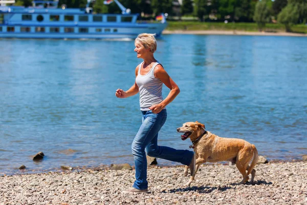 Reife Frau joggt mit Hund am Flussufer — Stockfoto