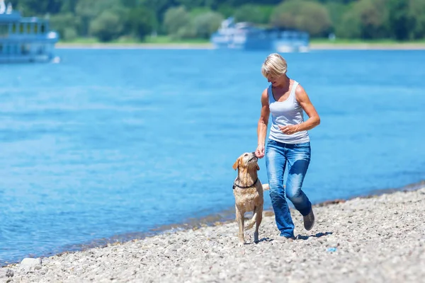 Rijpe vrouw jogs met een hond langs de rivier — Stockfoto