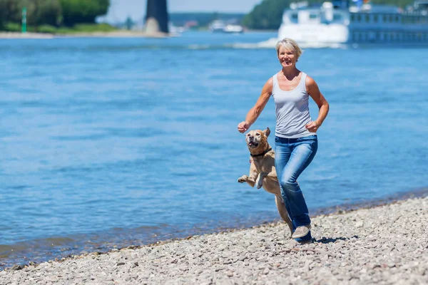 Reife Frau joggt mit Hund am Flussufer — Stockfoto