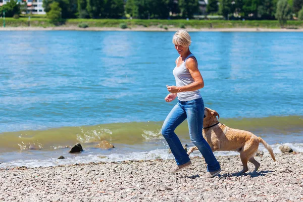 Reife Frau joggt mit Hund am Flussufer — Stockfoto