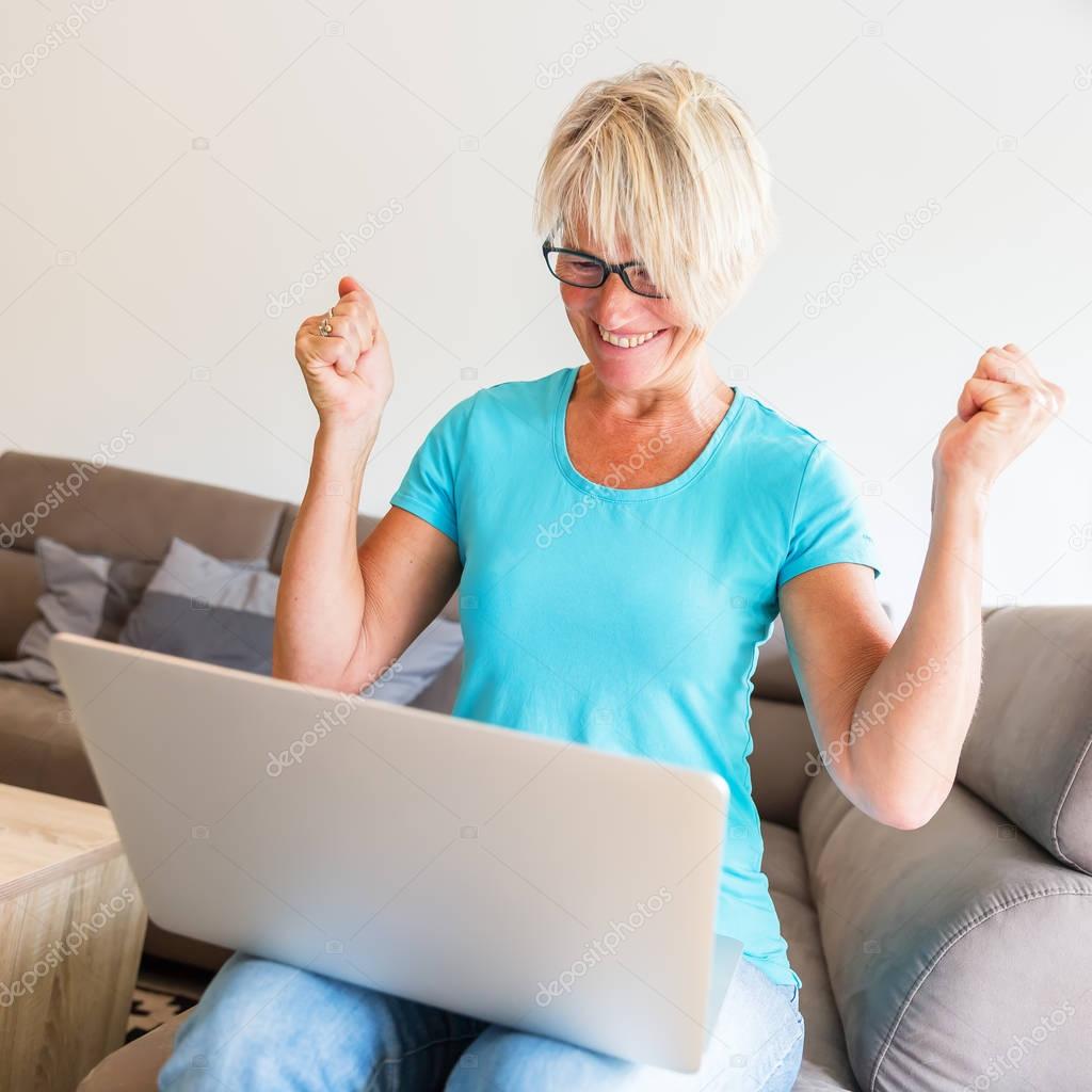 mature woman sits who is rejoicing with raised hands in front of a laptop