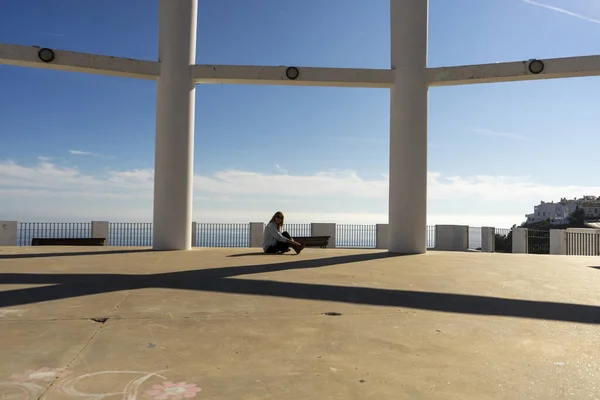 woman sitting on ground between columns and blue sea in background