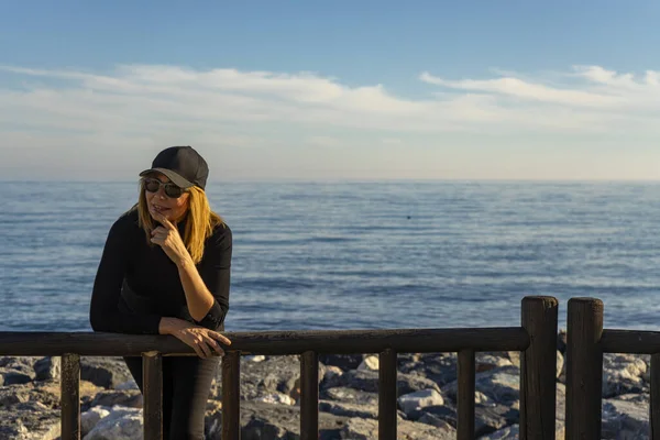 50歳以上のブロンドの女性はパンツと黒のシャツを着て帽子をかぶって海を背にポーズをとって ロイヤリティフリーのストック写真
