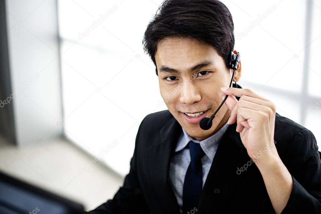 Happy confident smiling Asian business man staff at service desk talking on phone in a call centre, portrait of male operator agent with headsets, customer service or technical support concept