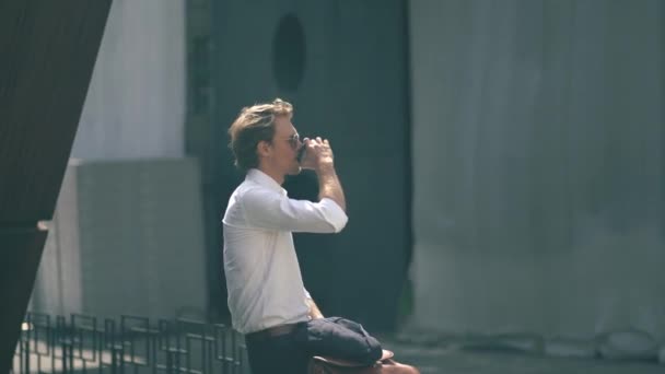 Bonito jovem bebendo café sentado no corrimão de metal — Vídeo de Stock