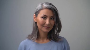 Gri arka planda açık mavi kazaklı, orta yaşlı Asyalı ya da Moğol bir kadın poz veriyor. Bayan nazikçe gülümsüyor ve diğer tarafa bakıyor. Yakın çekim.