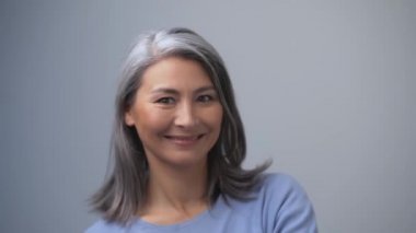 Çekici Asyalı kadın elleri havada komik bir yüz ifadesi takınıyor. Gri saçlarına dokunuyor. Gri arkaplandaki portre.