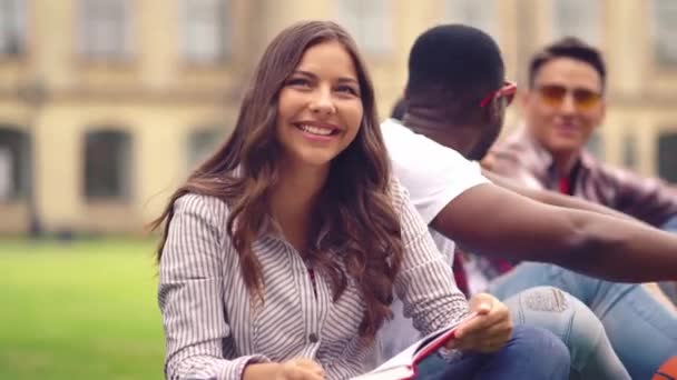 Student meisje lezen boek en glimlach zitten op het gras met haar vrienden — Stockvideo