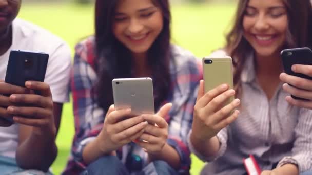 Jóvenes sentados y loughing mirando a un grupo de chat en sus teléfonos inteligentes — Vídeos de Stock