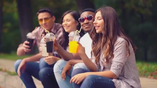 Los estudiantes felices se relajan sentados en el parque con jugos frescos — Vídeo de stock