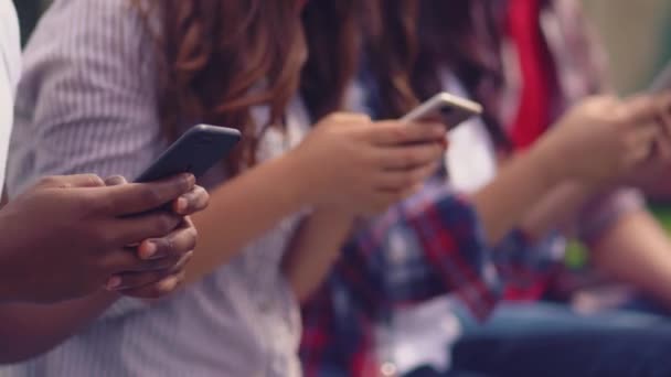 Los estudiantes que utilizan sus teléfonos inteligentes sentados al aire libre en una hierba verde . — Vídeo de stock