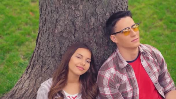 Two young students sitting leaned on a tree in park — Stock Video