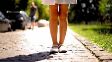 Parke taşının kenarında duran bir kızın ayaklarına yakın. Arkasında bir sürü araba olan ayakkabılarıyla yürüyen bir kız. Küçük bir kız hareketsiz duruyor.